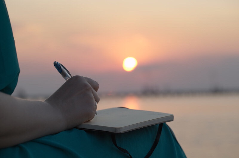 A woman’s hand writing in a diary with the ocean and sunset in the background.