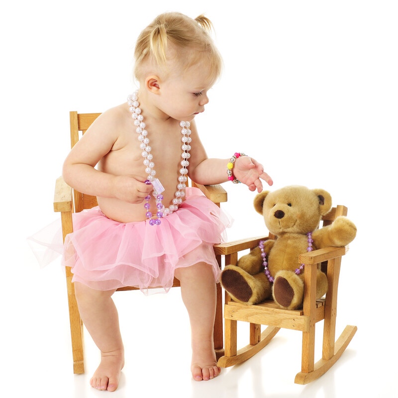 A toddler wearing beads and putting a necklace on her bear.